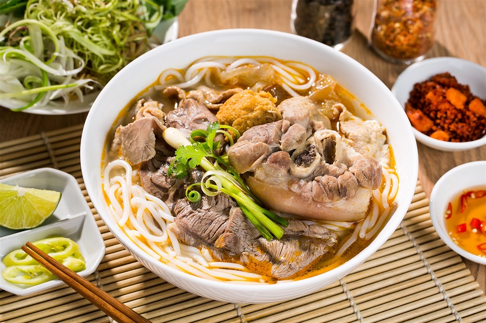 Bát bún bò đầy ắp giò ninh mềm, đậu hũ, rau thơm đặt cạnh bát muối chanh, đĩa rau sống