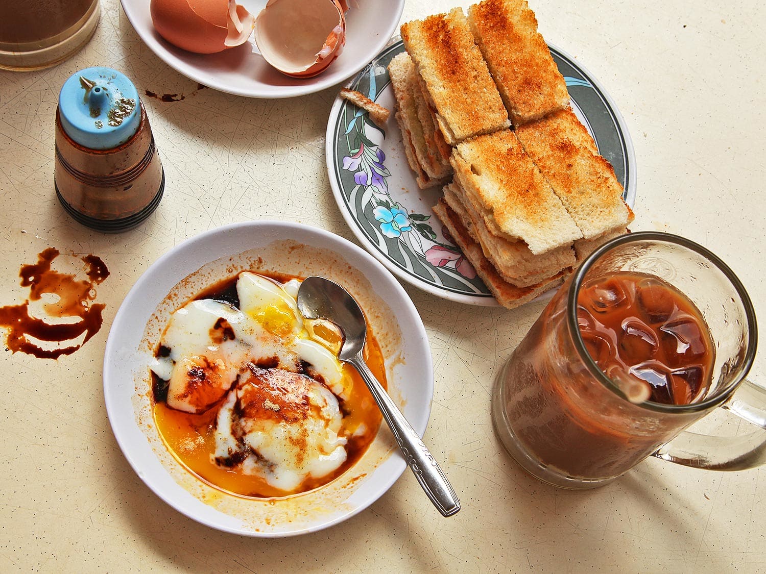 Đĩa bánh mỳ nướng vàng giòn phết bơ lạnh và mứt kaya, bên cạnh bát trứng luộc mềm và cốc cafe trên bàn