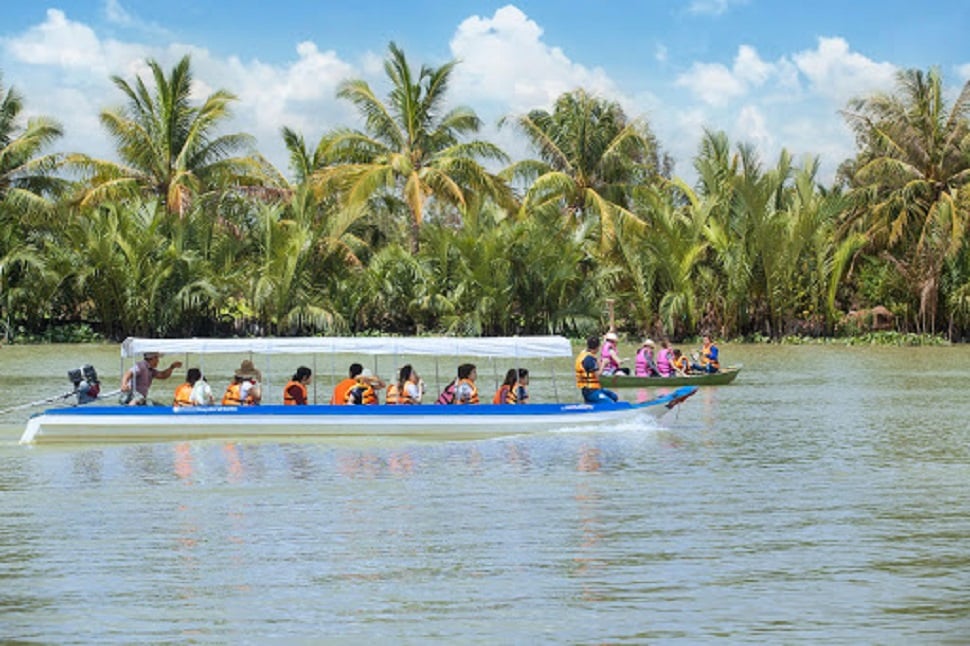 Nhóm du khách mặc áo phao ngồi thuyền chèo dọc con sông, với những hàng cây xanh mát