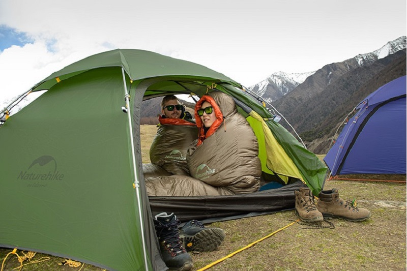 Đôi nam nữ sử dụng túi ngủ ngồi trong lều khi cắm trại trên đỉnh núi vào mùa đông