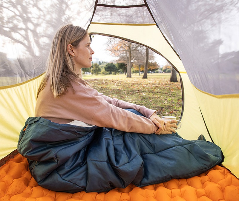 Người phụ nữ ngồi quấn trong túi ngủ xanh than trải trên đệm hơi trong lều
