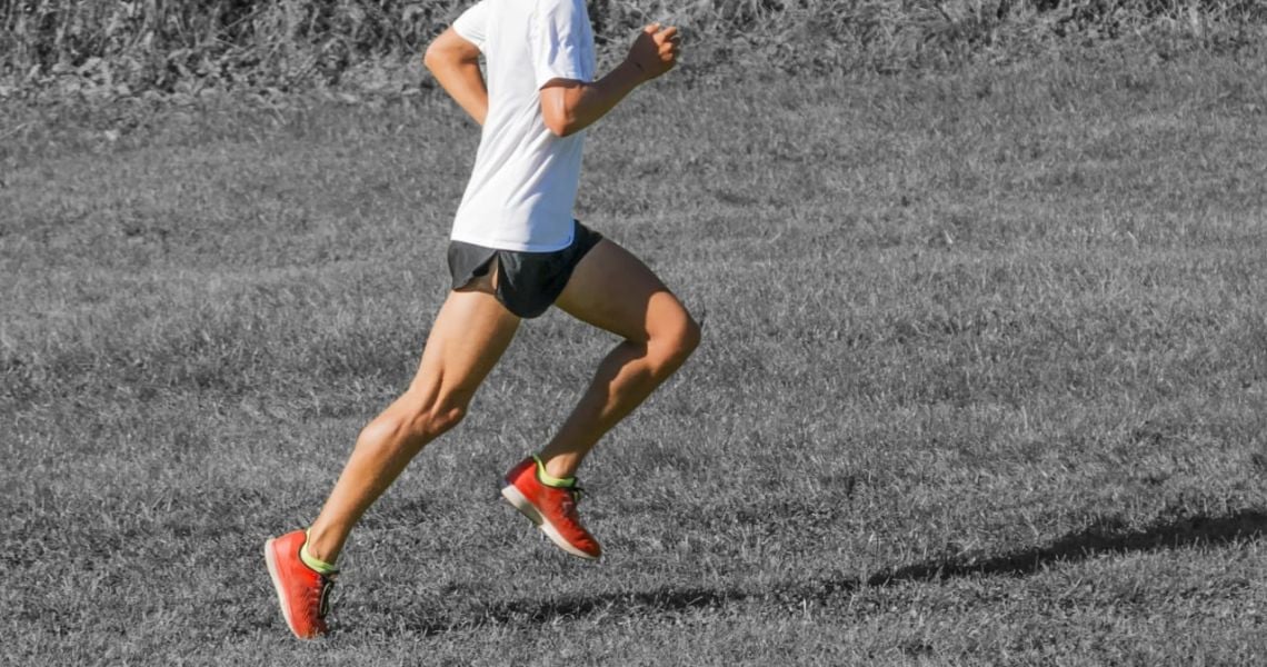 Người đang chạy trên bãi cỏ, mặc áo trắng và quần đen ngắn, mang giày thể thao màu đỏ.