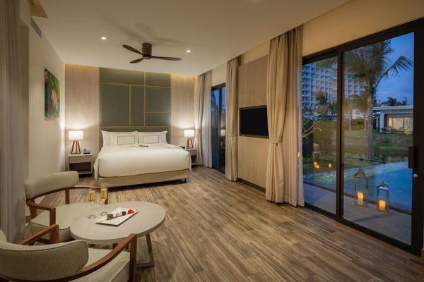 căn phòng có giường đôi, bàn ghế, sàn lót gỗ, và cửa lớn view ra biển vào buổi tối