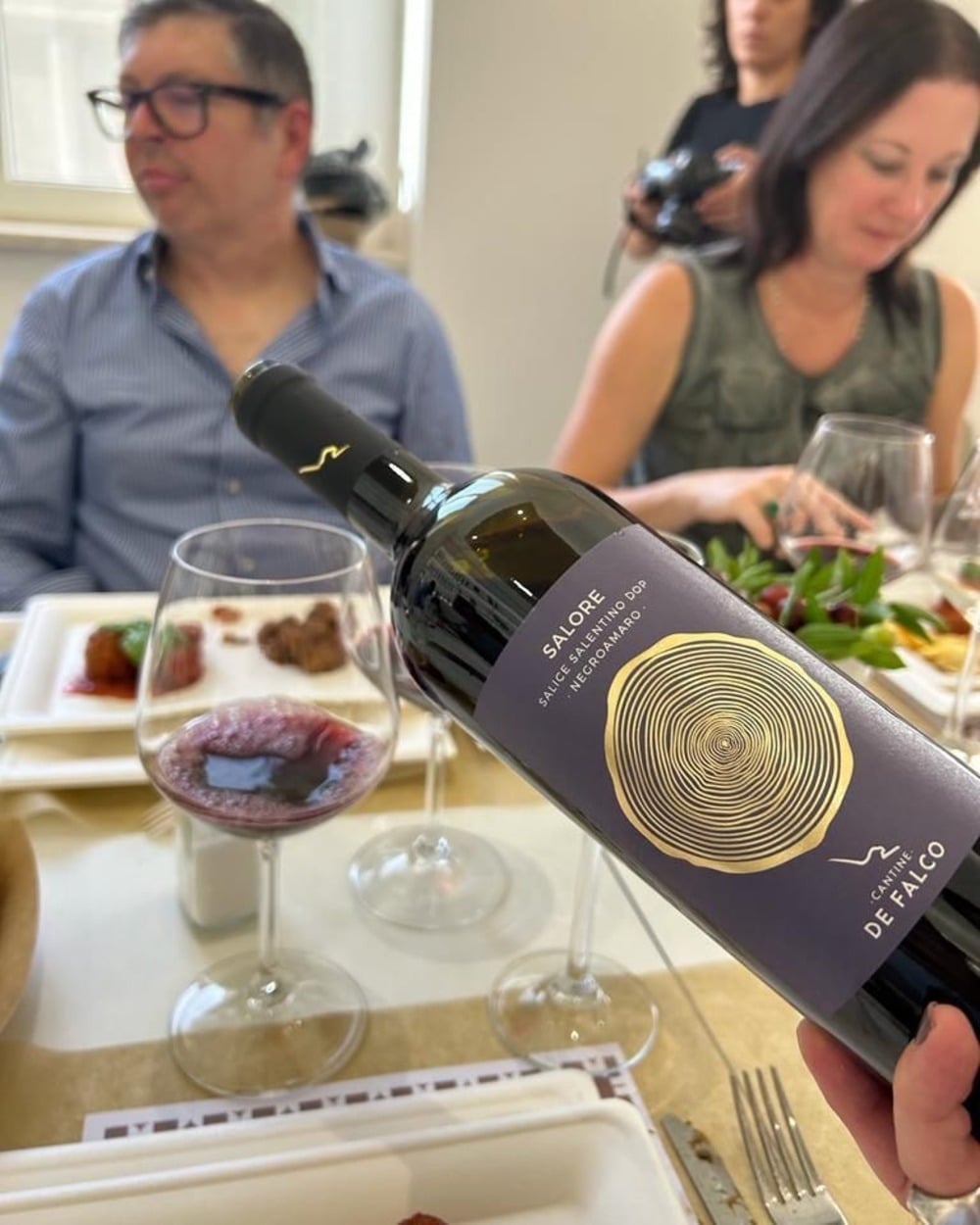 Ly rượu kiểu Bordeaux bên cạnh chai Negroamaro, minh họa phục vụ ở 16–18°C.
